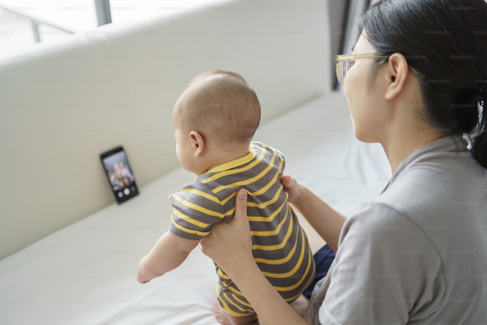 Happy Asian Young mother and newborn baby boy doing online video call with family on smartphone on bed at home. Mom and son using internet technology communication on mobile phone.