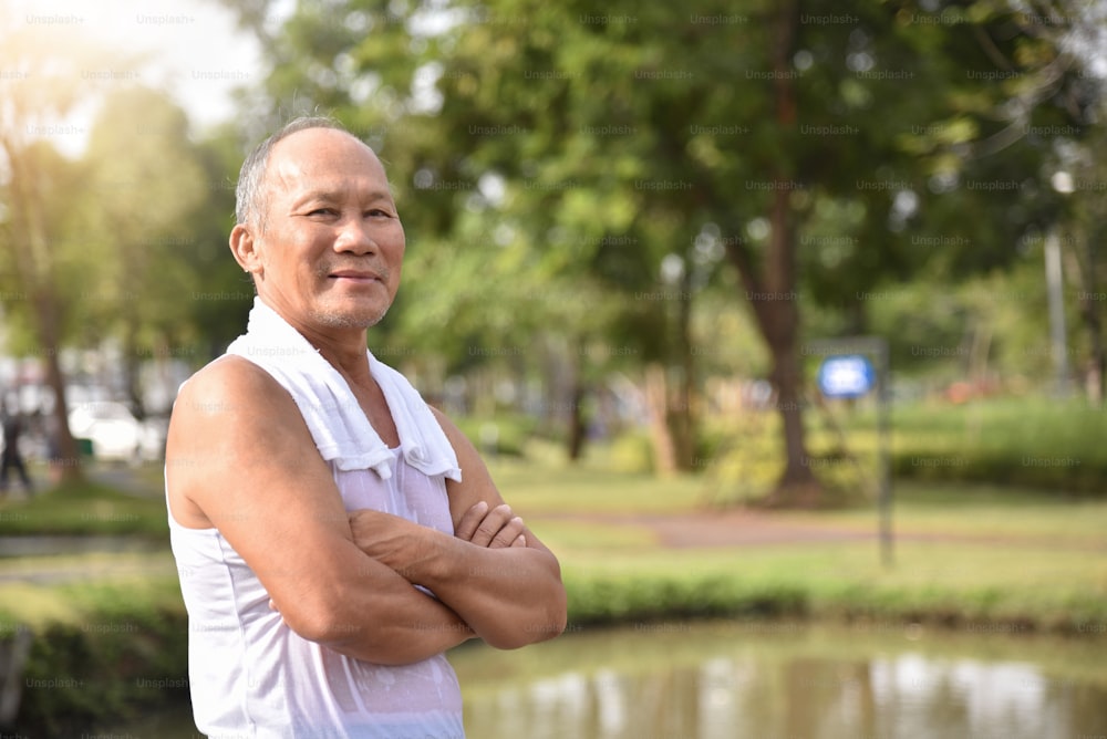 Maschio anziano asiatico fiducioso in posa con le braccia incrociate e sorridente mentre si esercita allo sfondo all'aperto del parco.