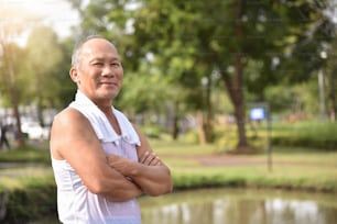 Selbstbewusster asiatischer älterer Mann, der mit verschränkten Armen posiert und lächelt, während er im Park im Hintergrund trainiert.