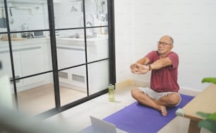 Homem adulto maduro asiático usando óculos sentado no tapete de ioga esticando os braços após o exercício em casa