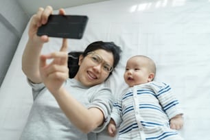 幸せな笑顔のアジアの若い母親と生まれたばかりの赤ちゃんがベッドに横たわって、自宅で一緒に携帯電話で自分撮りをしています。スマートフォンで楽しんでいる陽気なママと息子