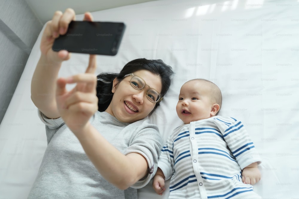 행복한 미소 짓는 아시아 젊은 엄마와 갓 태어난 아기가 침대에 누워 집에서 함께 휴대폰으로 셀카를 찍고 있다. 쾌활한 엄마와 아들이 스마트폰으로 즐거운 시간을 보내고 있다