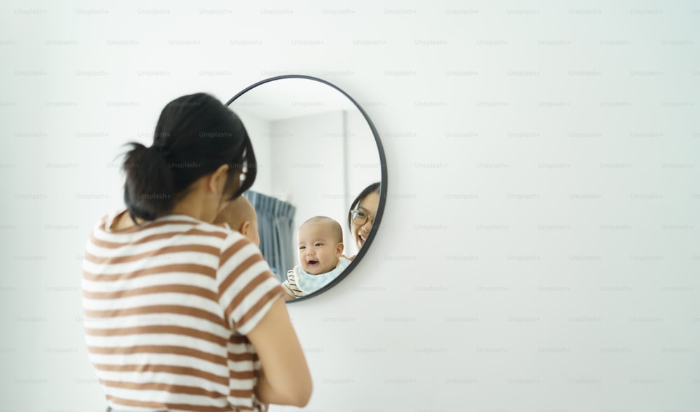 Smiling Asian mother holding little baby boy looking at mirror indoors
