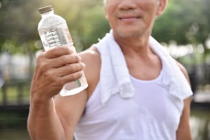 Hombre mayor asiático sosteniendo una botella de agua para beber mientras hace ejercicio en el fondo al aire libre del parque.