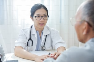 Young Asian Female doctor reassuring senior male patient with holding hands in medical office.