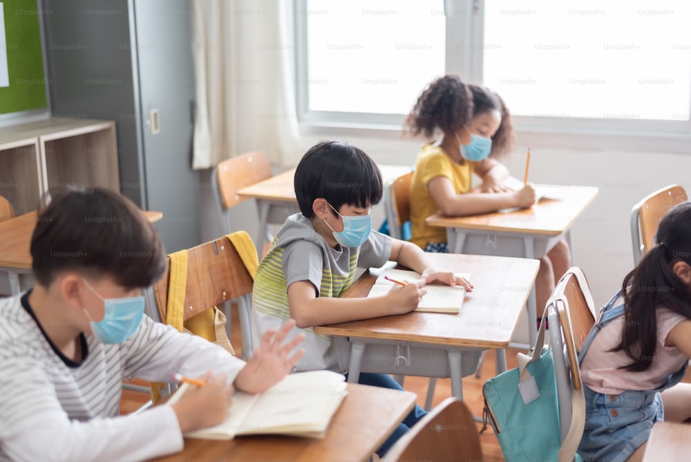 Concentrati Etnia diversa bambini delle scuole che indossano una maschera protettiva per il viso seduti al banco in classe, scrivendo.