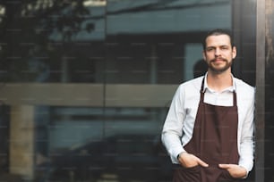 Successful SME. Male Barista Leaning against on front of the coffee shop glass wall. Portrait of Bearded man wear brown apron looking at camera with copy space.