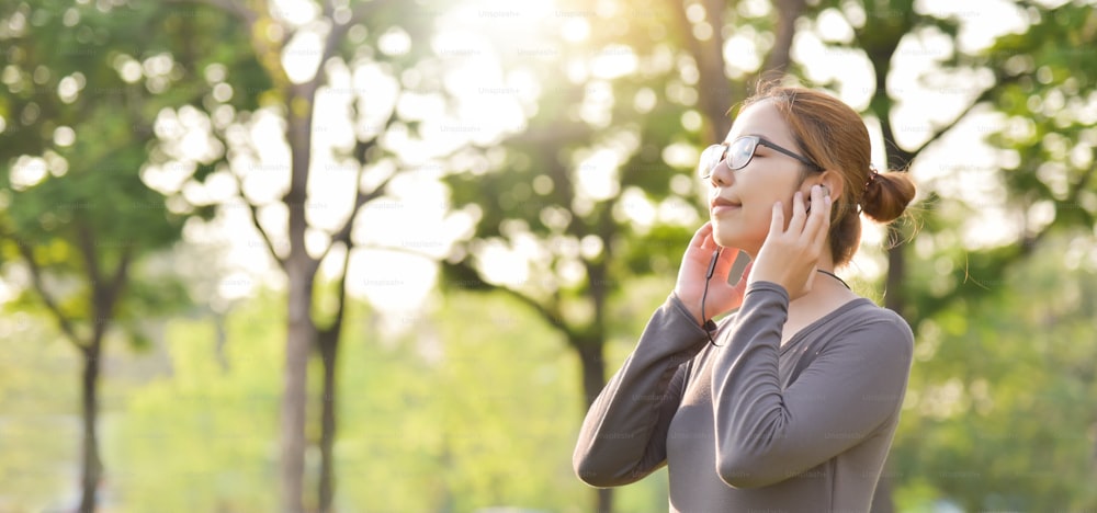 Happy Young Asian woman in Grey sportswear is smiling and listening to the music with earphones at outdoors on morning. Female workout at the park. Health care concept. Copy space.