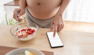Mujer embarazada usando el teléfono móvil mientras come alimentos saludables. Mujer asiática tocando la pantalla de un teléfono inteligente durante su embarazo en la cocina.