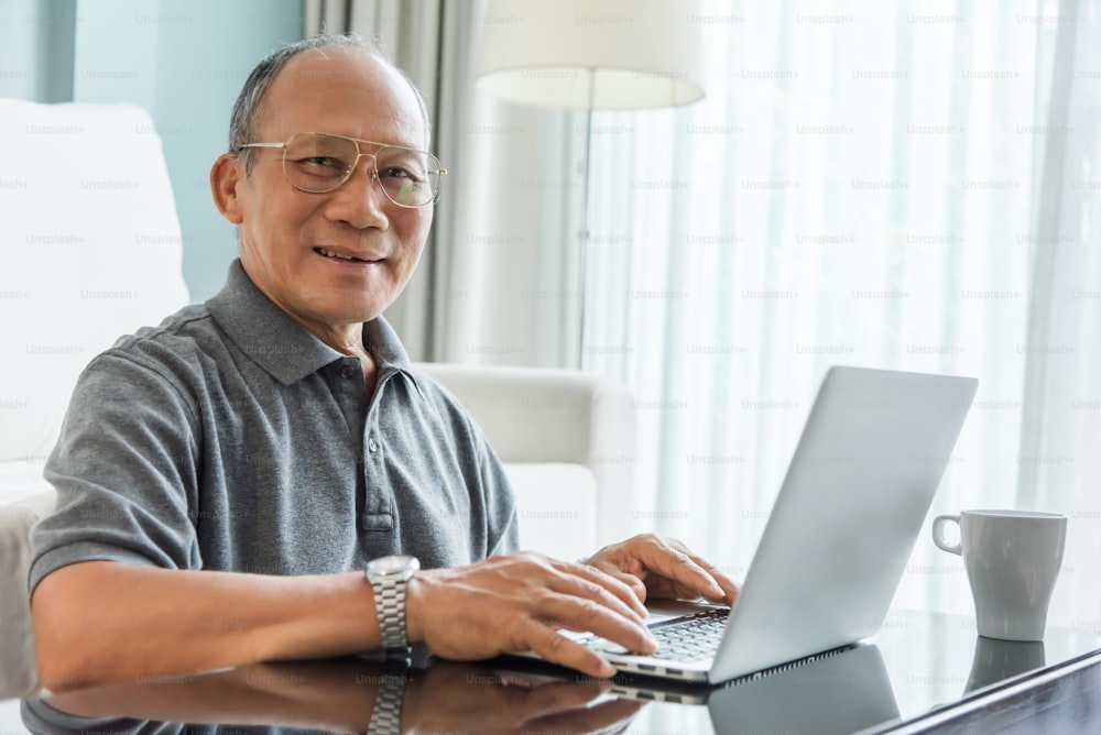 그의 집에서 노트북을 사용하는 편안한 아시아 노인. 행복한 노인 남성이 웃고 있다. 찾고 카메라. 퇴직