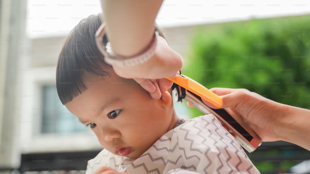 Menino pequeno asiático sendo cortado pelo cabelo por seus pais com cortador de cabelo ao ar livre
