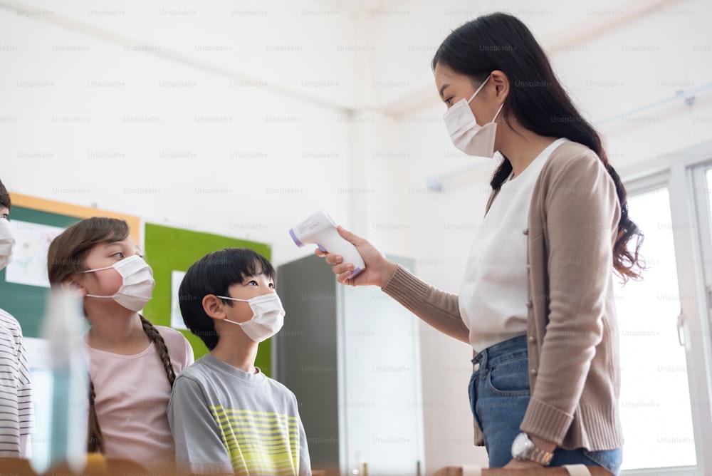 Young Teacher using digital thermometer for check temperature on Asian kid foreheads. Diversity students wearing protective face mask with COVID-19 Screening in the classroom at Elementary school