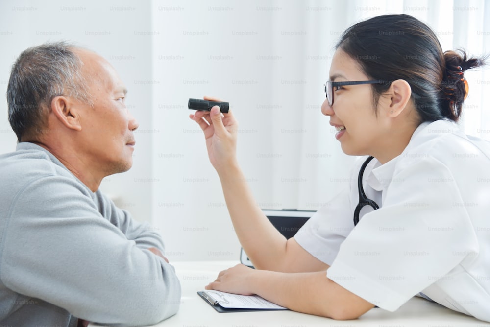若いアジアの女性医師は、診療所で懐中電灯で年配の男性の患者の目をチェックする眼鏡をかけています。