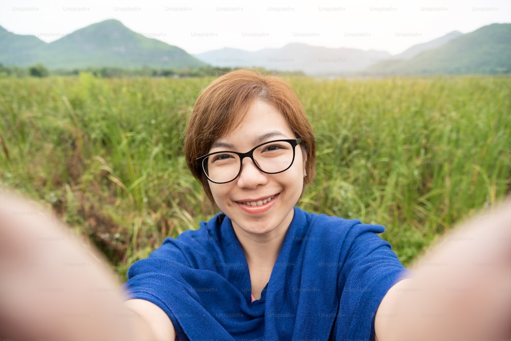 Happy Asian woman wear glasses taking a selfie with smartphone on field and mountain view. Smiling face.