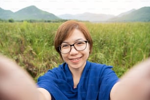 Happy Asian woman wear glasses taking a selfie with smartphone on field and mountain view. Smiling face.