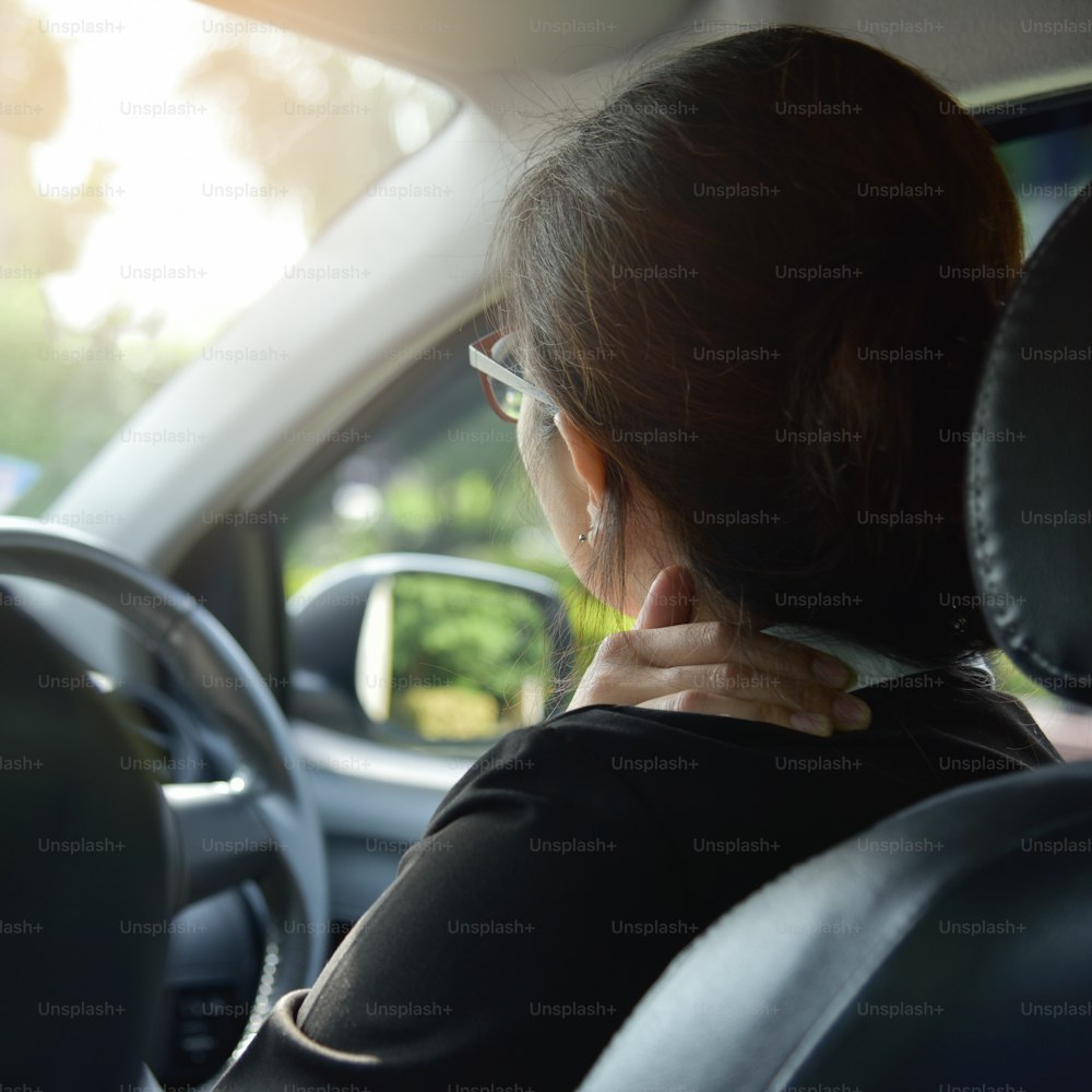 Vista posteriore della donna d'affari asiatica con occhiali che ha dolore alla spalla, alla schiena e al collo mentre guida un'auto. Malattia, esausto, malattia, stanco per il concetto di lavoro straordinario.