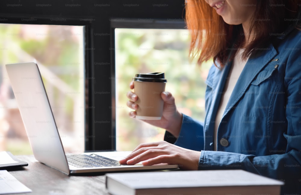 Bella donna asiatica con occhiali da lavoro in camicia blu seduta accanto alla finestra e con in mano una tazza di caffè mentre lavora con il computer portatile.