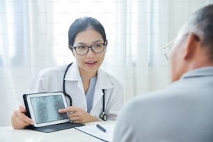 Jovem Médica Asiática usa óculos sorrindo e discutindo com o paciente do homem sênior sobre uma radiografia de tórax em tablet digital no consultório médico. Raio X.