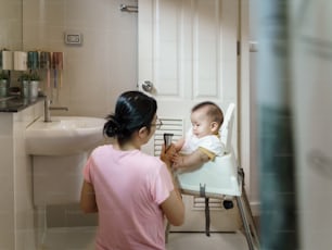 Asian Adorable little baby boy sitting on high chair having haircut in the bathroom with hair clipper by his Mother.