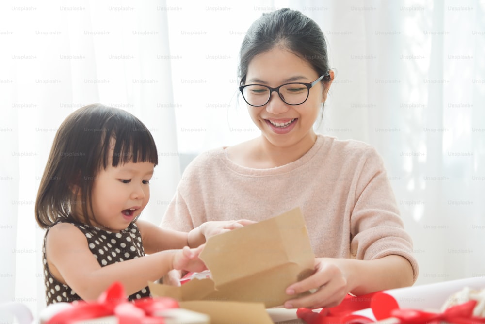 Giovane madre e sua figlia che confezionano una confezione regalo per compleanno, Natale e Capodanno in una stanza bianca. Felice famiglia asiatica a casa. Copia spazio.