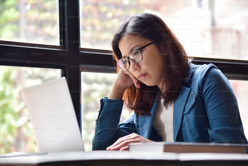 Donna asiatica stressata con occhiali in camicia blu che pensa e si siede accanto alla finestra mentre usa il laptop per il suo lavoro.