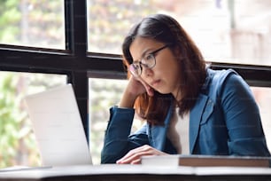 Mulher de óculos asiáticos estressados de camisa azul pensando e sentada ao lado da janela enquanto usava laptop para seu trabalho.