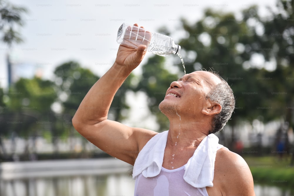 Homme senior asiatique versant de l’eau de la bouteille sur son visage après l’exercice dans le fond extérieur du parc. Lavage. Nettoyage.