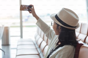 国際空�港での休暇中に携帯電話やスマートフォンで自撮りをしている笑顔の若いアジアの女性旅行者。