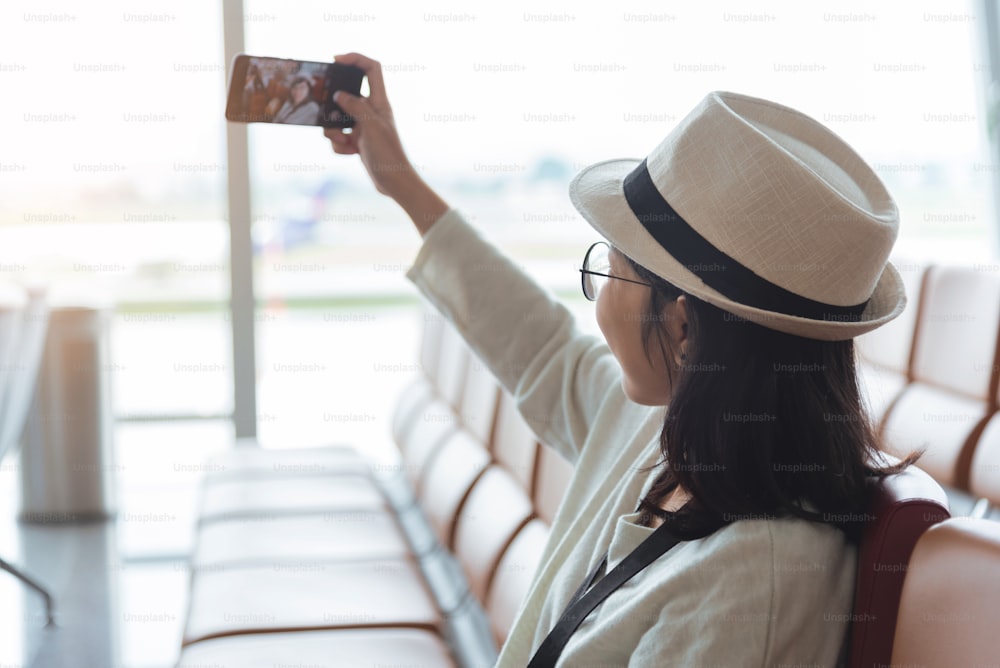 Smiling young Asian woman traveler taking a selfie with mobile phone or smartphone while travel in vacation at international airport.