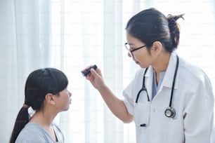 Young Asian Female Doctor wear glasses checking a little girl patient eyes with flashlight in medical office.