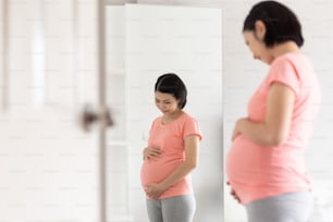 Cheerful Asian pregnant woman standing touching belly.