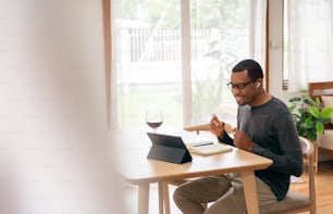 Heureux homme afro-américain dans des écouteurs utilisant une tablette numérique pour écouter de la musique et faire une célébration d’appel vidéo en ligne avec un toast au vin rouge à la maison