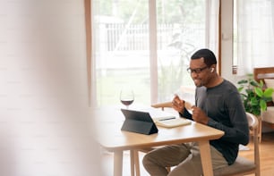 Happy African American man in earphones using digital tablet for listening to the music and making online video call celebration with toasting red wine at home