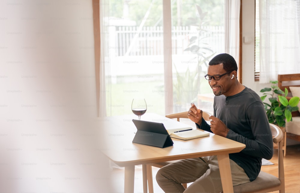 Happy African American man in earphones using digital tablet for listening to the music and making online video call celebration with toasting red wine at home