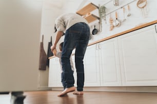 Glückliche asiatische Mutter, die Händchen ihres kleinen Kindes hält, das die ersten Schritte zu Hause macht, kleines Baby, das mit Mutterunterstützung in der Küche geht und läuft.