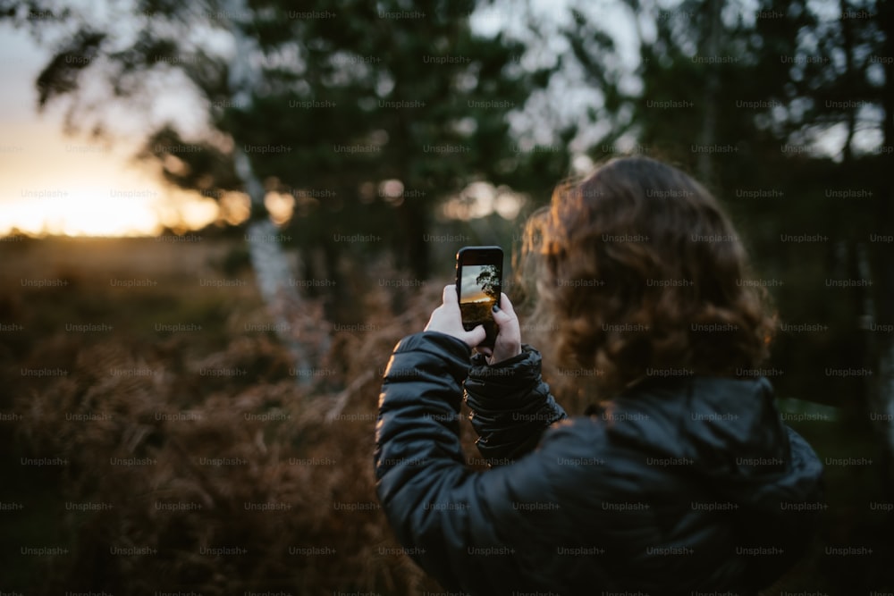 a woman taking a picture of the sun setting in the woods