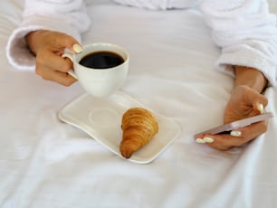 a woman sitting on a bed holding a cup of coffee and a croissant