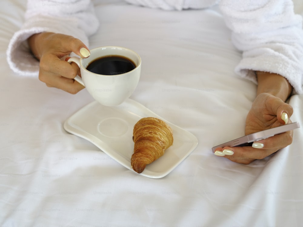 a woman sitting on a bed holding a cup of coffee and a croissant