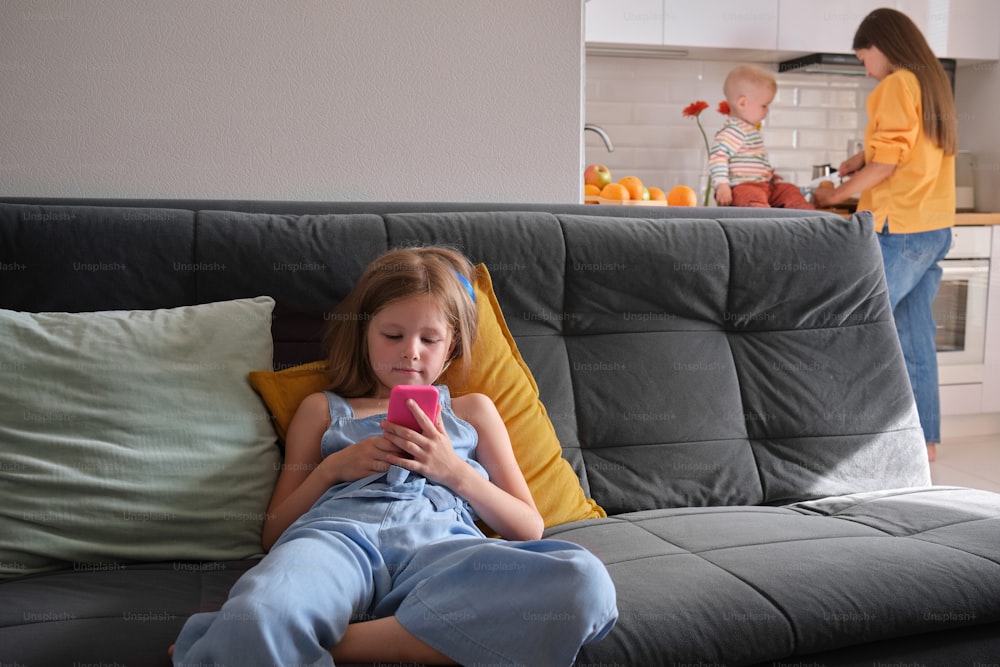 a little girl sitting on a couch looking at a cell phone