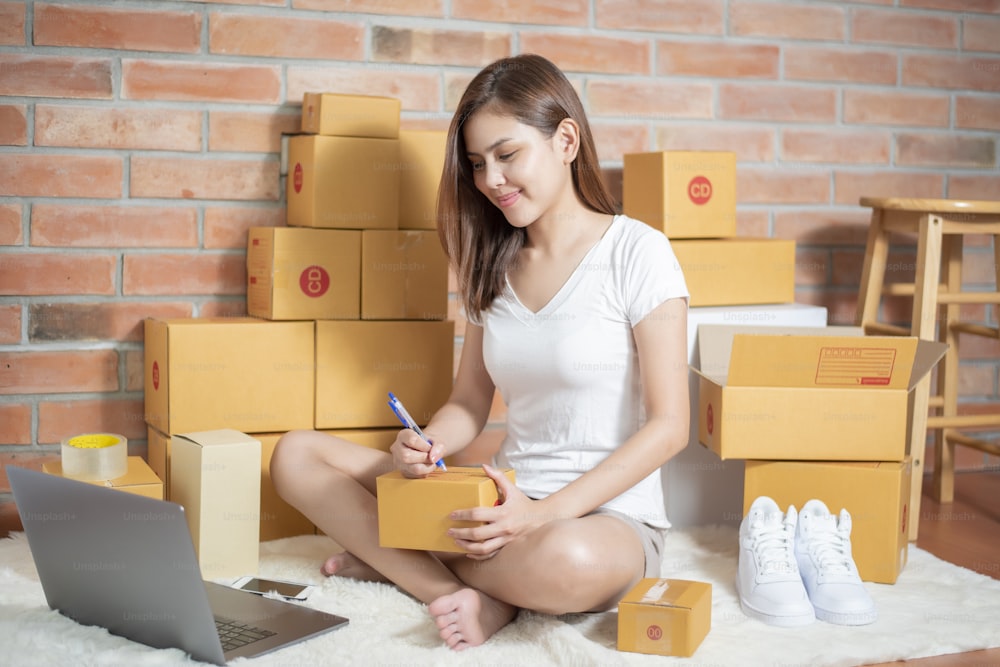 Woman entrepreneur owner SME business is checking order with smartphone, laptop and packaging box to send her customer