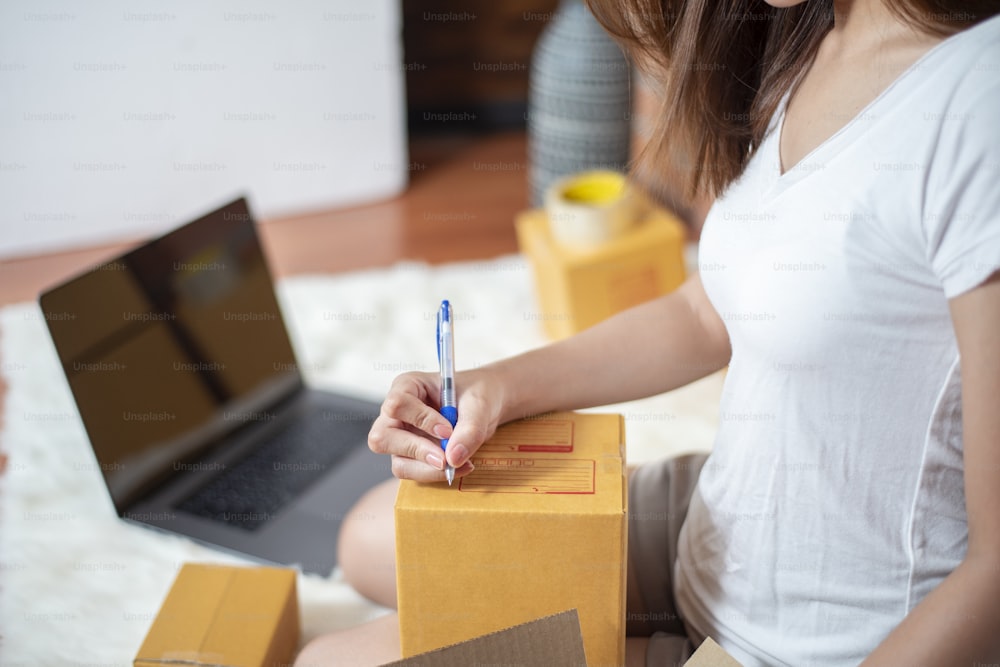 Une femme entrepreneure propriétaire d’une PME vérifie la commande avec un smartphone, un ordinateur portable et une boîte d’emballage à envoyer à son client