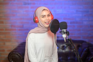 A young smiling muslim female singer wearing headphones with a microphone while recording song in a music studio with colorful lights.