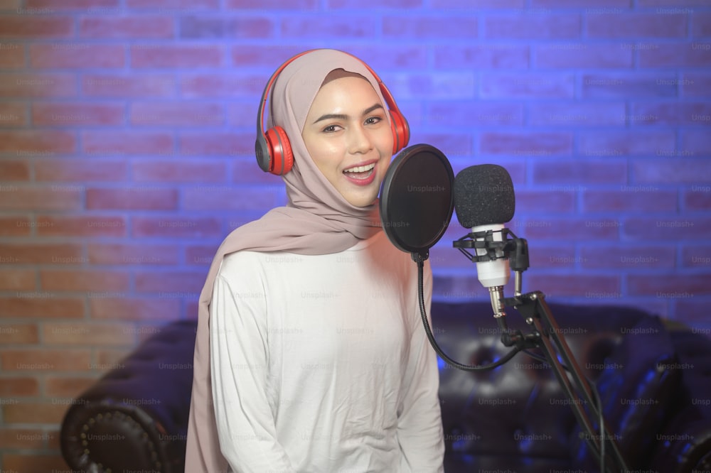 A young smiling muslim female singer wearing headphones with a microphone while recording song in a music studio with colorful lights.