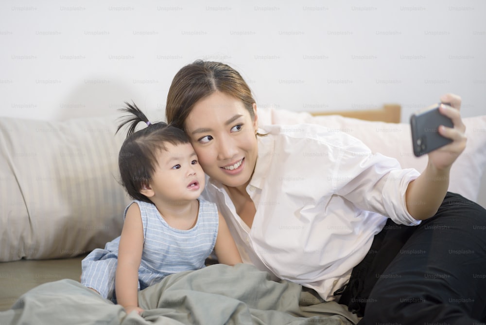 Asiatische Mutter und ihre kleine Tochter machen Selfie oder Videoanruf zum Vater im Bett, Familie, Haussicherheit, Elternschaft, Technologiekonzept.
