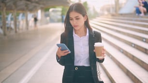Una giovane donna d'affari che indossa un vestito nero sta usando lo smartphone, tenendo una tazza di caffè in città, concetto di stile di vita aziendale