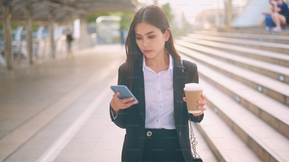 Uma jovem mulher de negócios vestindo terno preto está usando o telefone inteligente, segurando uma xícara de café na cidade, Business Lifestyle Concept