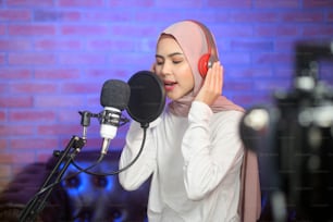 A young smiling muslim female singer wearing headphones with a microphone while recording song in a music studio with colorful lights.