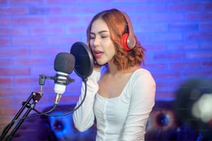 A young smiling female singer wearing headphones with a microphone while recording song in a music studio with colorful lights.