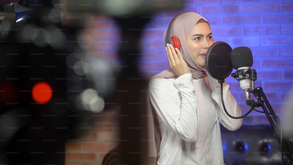 Una joven cantante musulmana sonriente con auriculares con micrófono mientras graba una canción en un estudio de música con luces de colores.