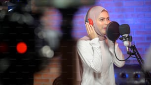 A young smiling muslim female singer wearing headphones with a microphone while recording song in a music studio with colorful lights.